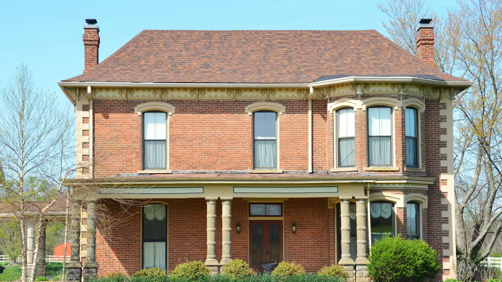 A large brick house with a deep covered porch set back behind mature trees on a residential street