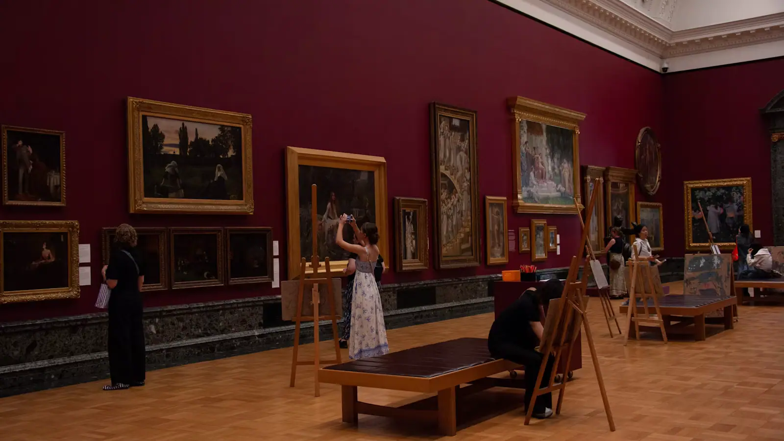 Two people standing back from a row of framed paintings hung on a white gallery wall