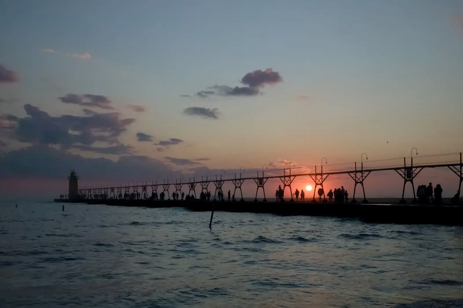 Silhouetted figures walking a long concrete pier toward a lighthouse under low sun
