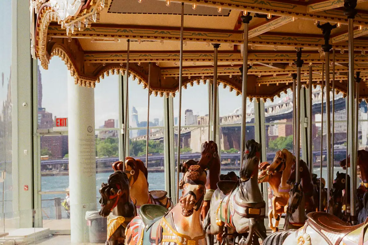Ornately painted carousel horses standing inside a glass pavilion with a city view beyond