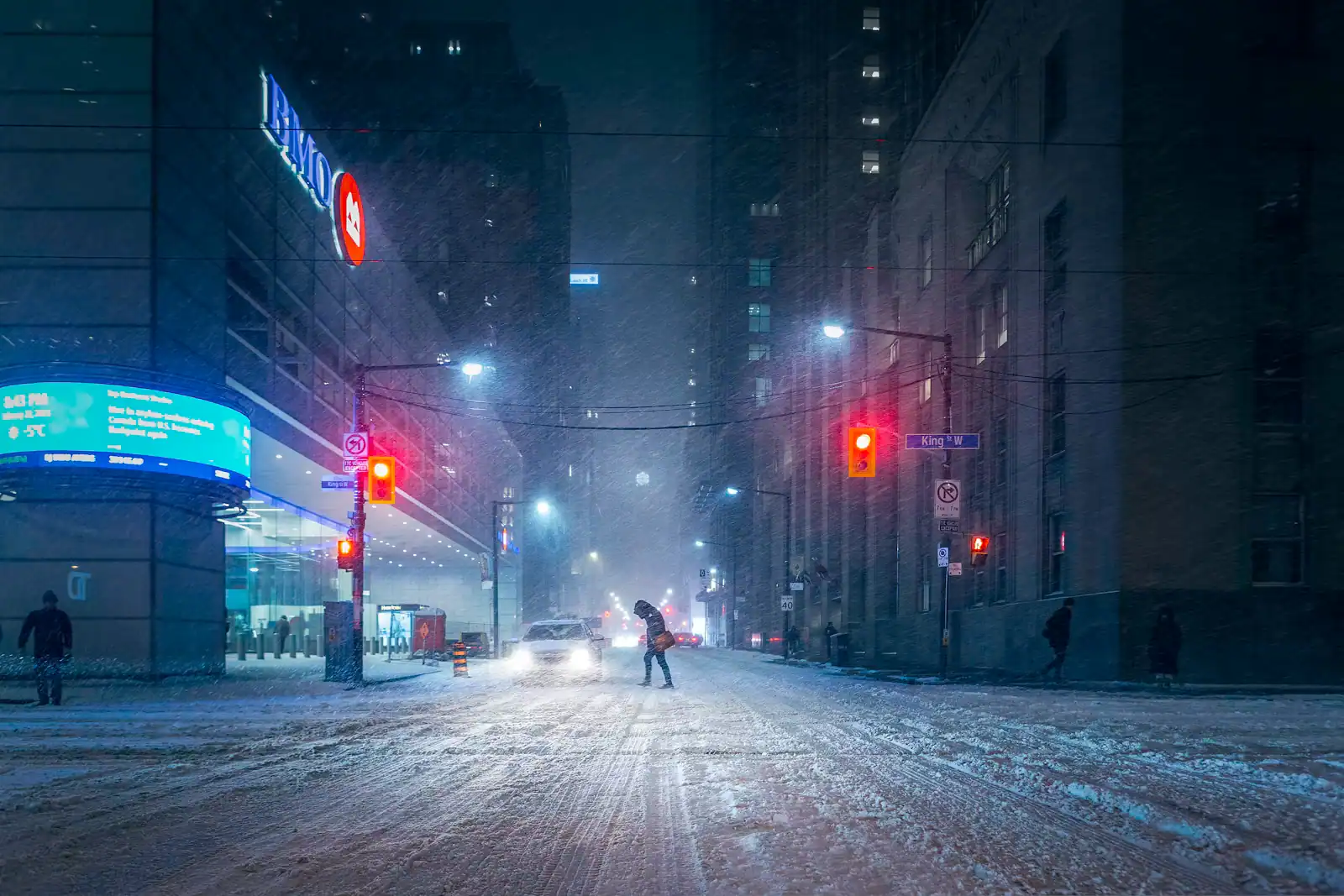 Two people crossing a snow-covered city street between parked cars